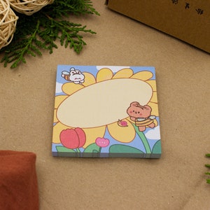 Peut inclure: Un tournesol jaune avec un ours brun portant un costume d'abeille et une abeille blanche volant au-dessus. Le tournesol a un espace vide au centre pour écrire. L'arrière-plan est bleu avec des fleurs roses et des feuilles vertes.