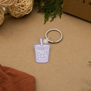 Peut inclure: Un porte-clés en acrylique transparent avec un contour violet d'un gobelet de boba tea avec une paille et des perles de boba à l'intérieur. Le porte-clés a un anneau en métal argenté.