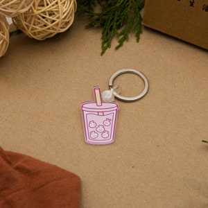 Peut inclure: Un porte-clés en forme de bubble tea rose et blanc avec une paille et un anneau de clé argenté. Le porte-clés représente une illustration de dessin animé d'un verre de bubble tea avec des perles de boba.