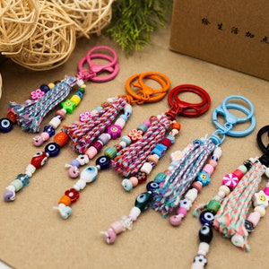 Peut inclure: Six porte-clés colorés avec des glands et des perles. Chaque porte-clés est doté d'un anneau métallique de couleur différente et d'une variété de perles colorées, notamment des perles bleues en forme d'œil maléfique, des perles roses, blanches et bleues, ainsi que de petits breloques colorés. Les porte-clés sont disposés sur une surface brune.