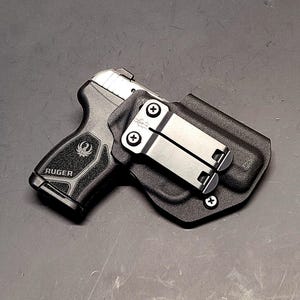 May include: A black Ruger handgun in a black holster with a silver metal clip. The handgun is black with a silver slide and the word "Ruger" is visible on the grip. The holster has a textured surface.