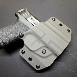 May include: A black handgun with a textured grip is holstered in a gray leather holster with multiple black screws.