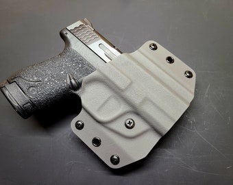Puede incluir: Una pistola negra con un agarre texturizado está enfundada en una funda de cuero gris con varios tornillos negros.