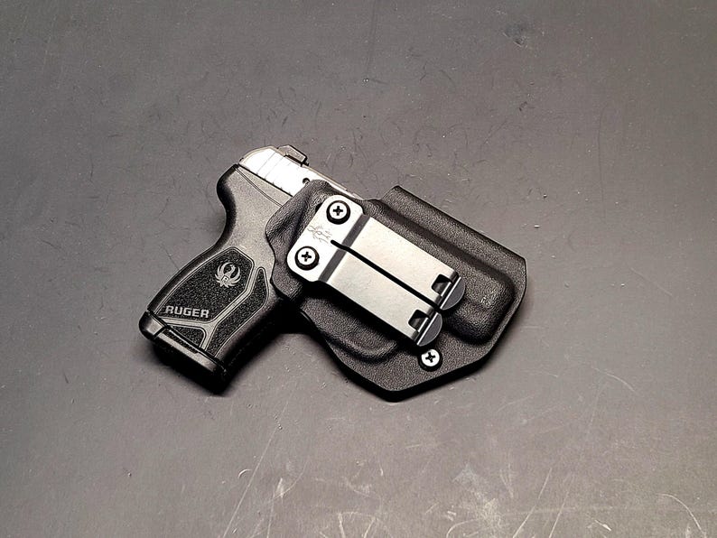 May include: A black Ruger handgun in a black holster with a silver metal clip. The handgun is black with a silver slide and the word "Ruger" is visible on the grip. The holster has a textured surface.