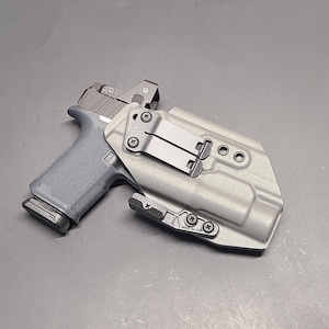 May include: A gray handgun with a red dot sight is placed inside a light gray holster. The holster has a metal clip and is secured with screws. The gun is positioned at an angle, showcasing its design and features.