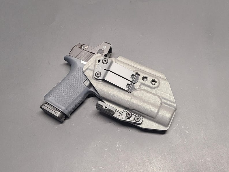 May include: A gray handgun with a red dot sight is placed inside a light gray holster. The holster has a metal clip and is secured with screws. The gun is positioned at an angle, showcasing its design and features.