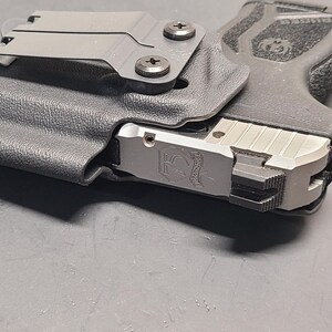 May include: A black polymer holster with a silver firearm. The holster has a metal clip and is secured with two screws. The firearm has a silver slide with engraved details and a black grip.