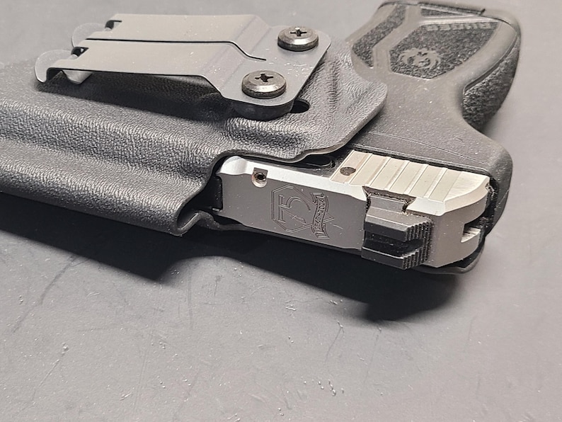 May include: A black polymer holster with a silver firearm. The holster has a metal clip and is secured with two screws. The firearm has a silver slide with engraved details and a black grip.