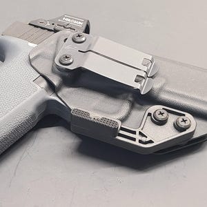 May include: A black polymer holster with a grey handgun. The holster has a metal clip and is secured with black screws. The handgun has a textured grip and a red dot sight.