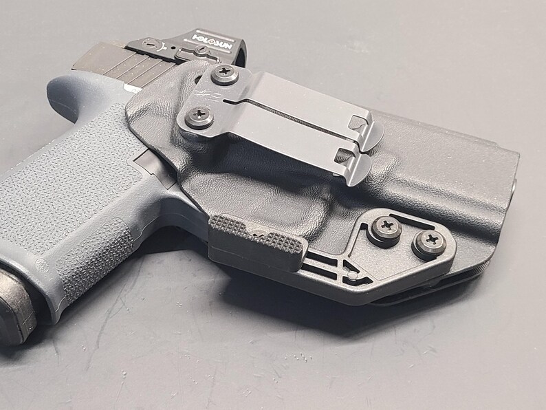 May include: A black polymer holster with a grey handgun. The holster has a metal clip and is secured with black screws. The handgun has a textured grip and a red dot sight.