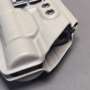 May include: A light gray, textured holster with a black firearm barrel visible. The holster has metal hardware and is designed to securely hold a handgun. The design includes a belt clip and several rivets.