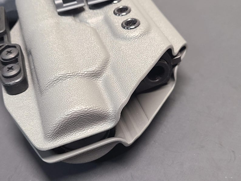 May include: A light gray, textured holster with a black firearm barrel visible. The holster has metal hardware and is designed to securely hold a handgun. The design includes a belt clip and several rivets.