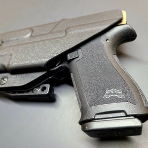 May include: A black polymer handgun with a black polymer holster. The handgun has a black polymer grip with a "PISTOLERA" logo. The holster has a black polymer clip.