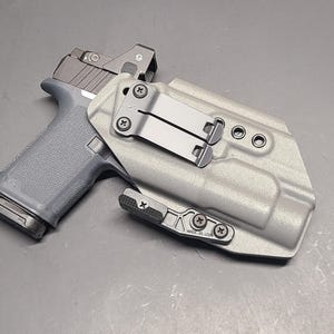 May include: A gray handgun with a red dot sight is placed inside a light gray holster. The holster has a metal clip and is secured with black screws. The handgun is gray with a black slide.