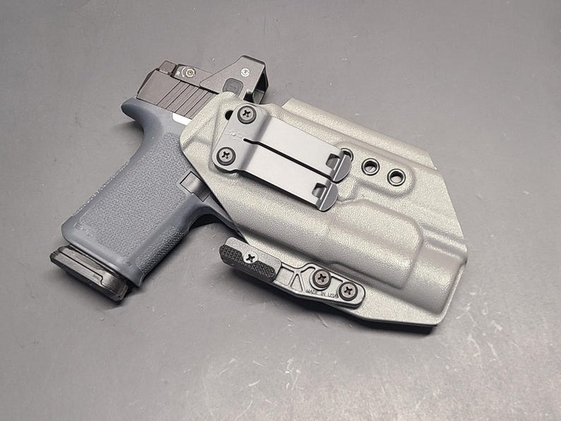 May include: A gray handgun with a red dot sight is placed inside a light gray holster. The holster has a metal clip and is secured with black screws. The handgun is gray with a black slide.
