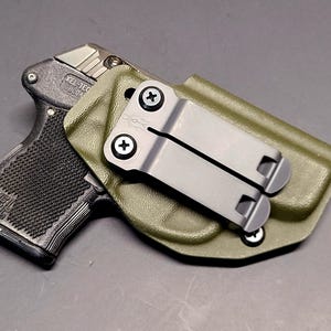 May include: A black handgun with a textured grip is partially inserted into an olive green holster. The holster has a metal clip and is secured with screws. The gun is marked with the text "KEL-TEC" and "COLOA".