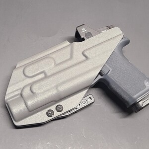 May include: A gray pistol with a red dot sight is partially inserted into a gray holster. The holster has a textured surface with molded details and is secured with black screws. The pistol grip is dark gray.