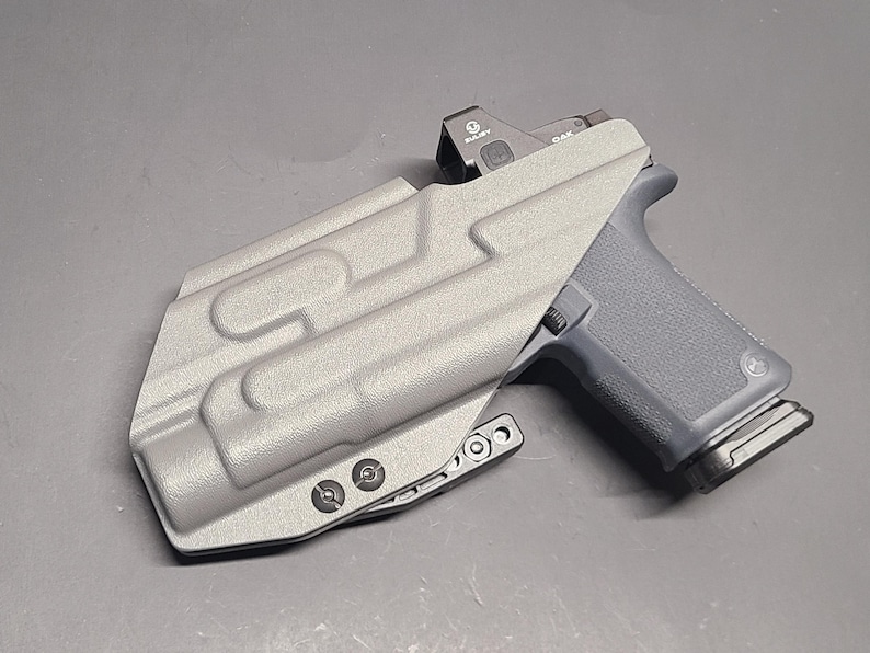 May include: A gray pistol with a red dot sight is partially inserted into a gray holster. The holster has a textured surface with molded details and is secured with black screws. The pistol grip is dark gray.