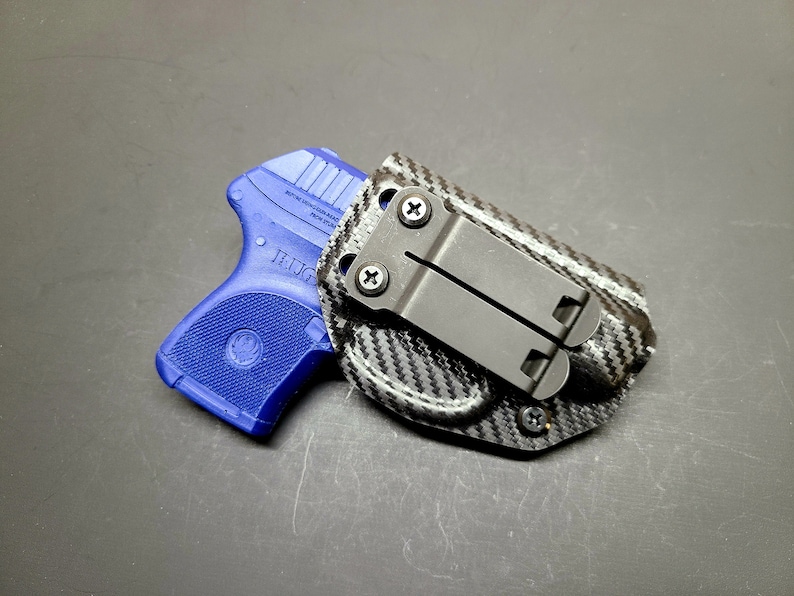 May include: A blue handgun with a black carbon fiber holster and a clip. The holster has a black metal clip with screws.