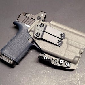 May include: A gray polymer holster with a black pistol. The pistol has a blue grip and a red dot sight. The holster features a black metal clip and is secured with black screws.