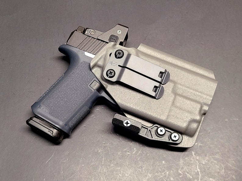 May include: A gray polymer holster with a black pistol. The pistol has a blue grip and a red dot sight. The holster features a black metal clip and is secured with black screws.
