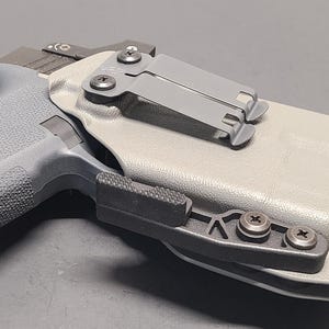 May include: A gray handgun in a light gray holster with a black clip. The holster has a black attachment with three screws. The handgun has a textured grip and a black slide.