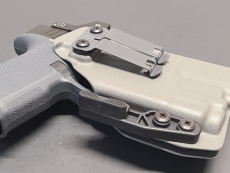 May include: A gray handgun in a light gray holster with a black clip. The holster has a black attachment with three screws. The handgun has a textured grip and a black slide.