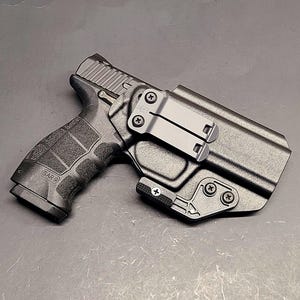 May include: A black SAR 9 handgun in a black holster. The handgun has a textured grip and the text "SAR 9" is visible. The holster has a metal clip and is secured with screws. The background is a solid gray.