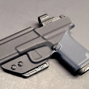 May include: A black polymer holster with a firearm. The firearm has a dark grey grip and a red dot sight. The holster is designed to securely hold the firearm. The image is taken from a slightly elevated angle, showcasing the details of the holster and firearm.