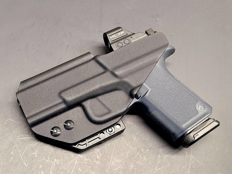 May include: A black polymer holster with a firearm. The firearm has a dark grey grip and a red dot sight. The holster is designed to securely hold the firearm. The image is taken from a slightly elevated angle, showcasing the details of the holster and firearm.