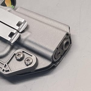 May include: A close-up of a grey, moulded polymer holster with a metal clip and screws. The holster is designed to securely hold a firearm. The metal clip is silver and has two square openings. The screws are silver and hold the clip in place.