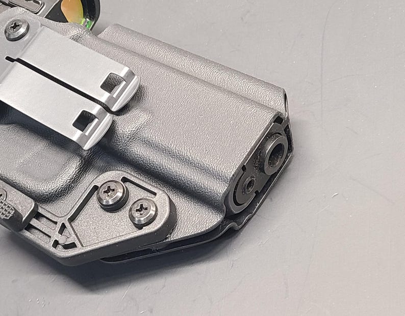 May include: A close-up of a grey, moulded polymer holster with a metal clip and screws. The holster is designed to securely hold a firearm. The metal clip is silver and has two square openings. The screws are silver and hold the clip in place.