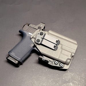May include: A black handgun with a gray slide and a red dot sight is secured in a gray holster. The holster has a black clip and is mounted on a dark surface. The gun's grip is a dark blue color.