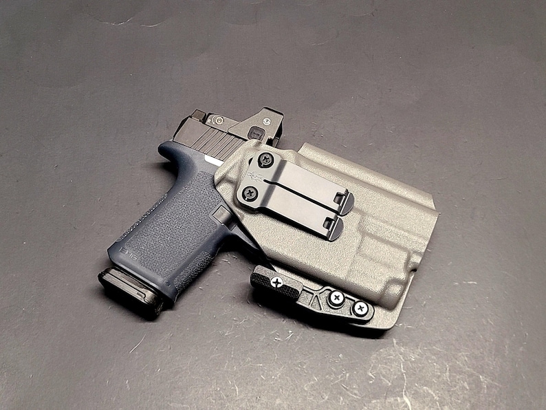 May include: A black handgun with a gray slide and a red dot sight is secured in a gray holster. The holster has a black clip and is mounted on a dark surface. The gun's grip is a dark blue color.
