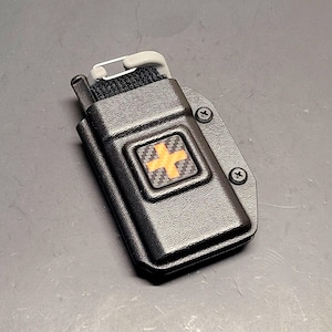 May include: Black tactical medical pouch with a prominent orange cross emblem. The pouch is secured with a black strap and features a gray safety pin. The design includes a textured surface and is mounted with black screws.