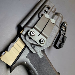 May include: A black handgun holster with a brown leather belt clip. The holster has a textured surface and a black plastic clip. The holster is designed to hold a handgun with a magazine.