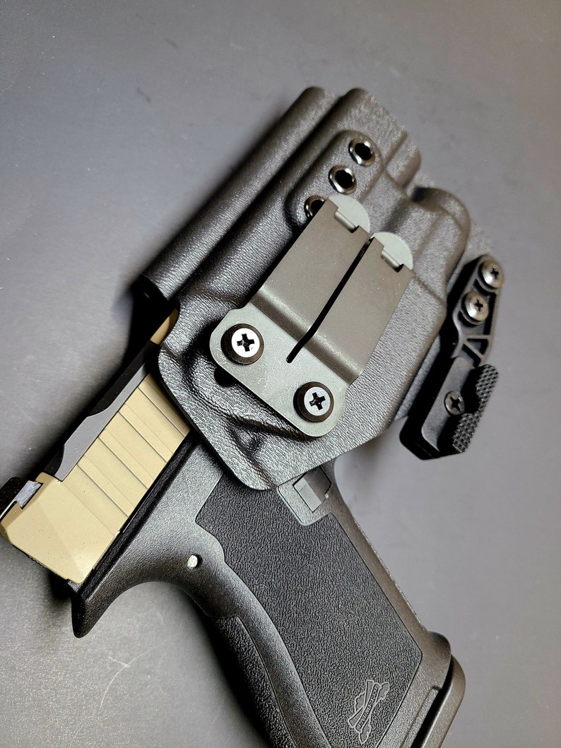 May include: A black handgun holster with a brown leather belt clip. The holster has a textured surface and a black plastic clip. The holster is designed to hold a handgun with a magazine.