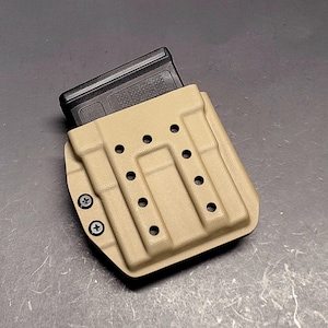 May include: Tan Kydex magazine carrier with a black magazine inserted. The carrier has multiple holes for mounting and is secured with black screws. The design is for carrying a magazine.