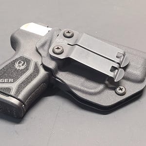 May include: A black Ruger handgun in a black holster. The handgun is visible with the Ruger logo and text. The holster has a metal clip for attachment. The image is a close-up shot.
