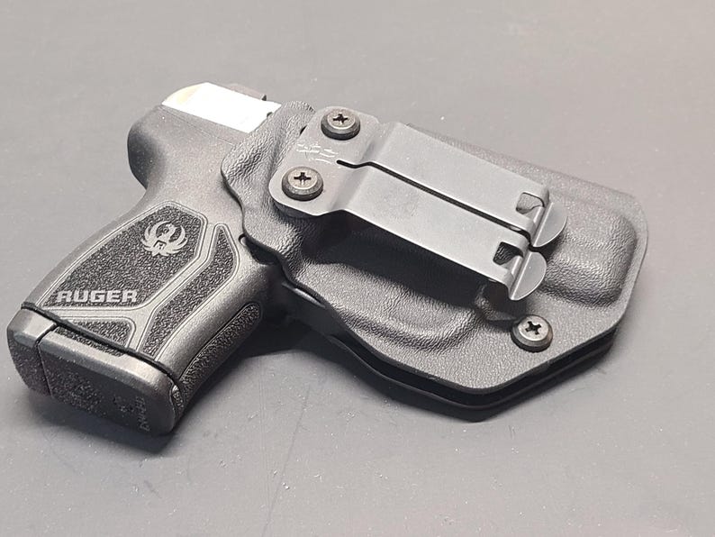 May include: A black Ruger handgun in a black holster. The handgun is visible with the Ruger logo and text. The holster has a metal clip for attachment. The image is a close-up shot.