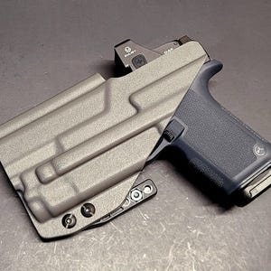 May include: A gray pistol in a matching gray holster. The pistol has a black grip and a red dot sight. The holster has a textured surface and is secured with screws.