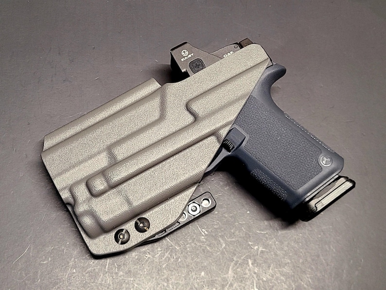 May include: A gray pistol in a matching gray holster. The pistol has a black grip and a red dot sight. The holster has a textured surface and is secured with screws.