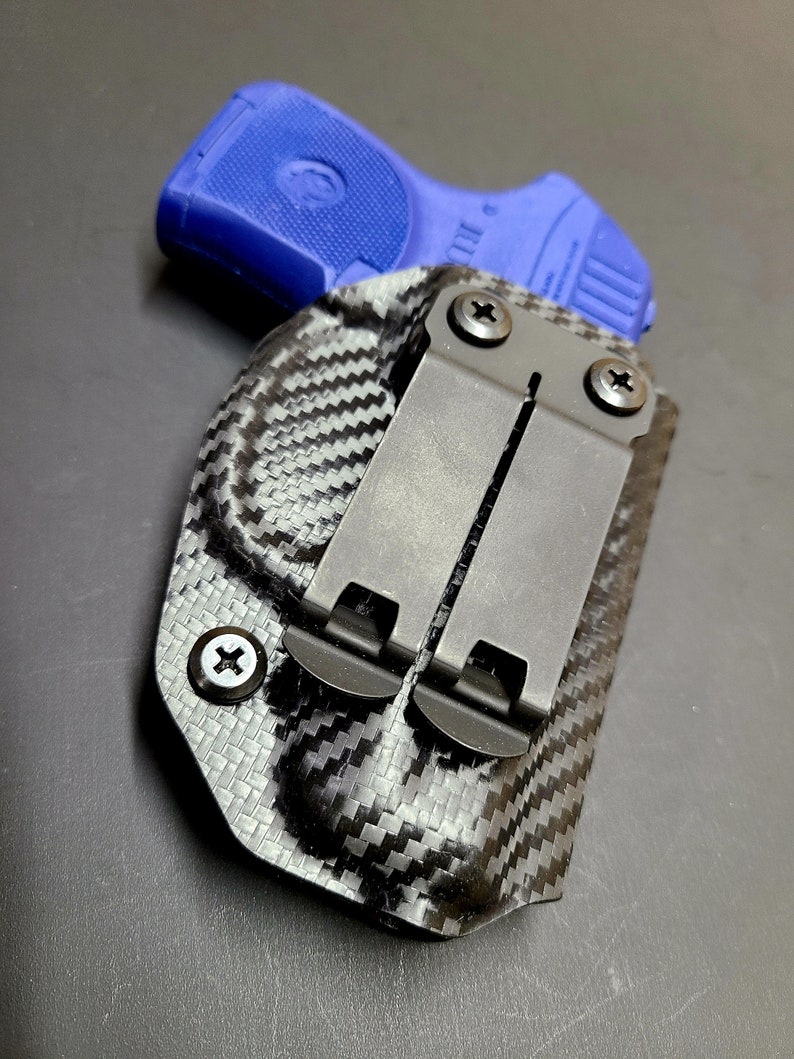 May include: A black carbon fiber holster with a blue polymer handgun inside. The holster has a metal clip with screws.