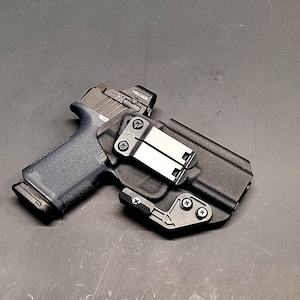 May include: A black handgun with a red dot sight is in a black holster. The holster has a metal clip and is secured with screws. The gun has a dark blue grip. The background is a dark grey surface.