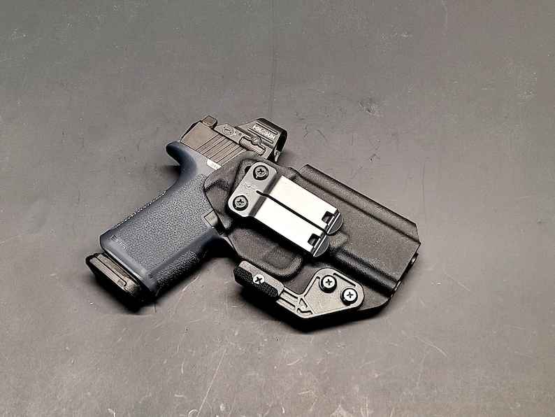 May include: A black handgun with a red dot sight is in a black holster. The holster has a metal clip and is secured with screws. The gun has a dark blue grip. The background is a dark grey surface.