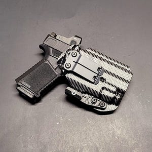 May include: A black handgun with a mounted optic sight is holstered in a patterned black and white holster. The holster has a metal clip for attachment. The firearm is set against a dark gray background.
