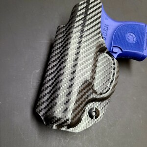 May include: A black carbon fiber holster for a small handgun. The holster has a single screw on the bottom and the text "CLEAR" is visible on the side.