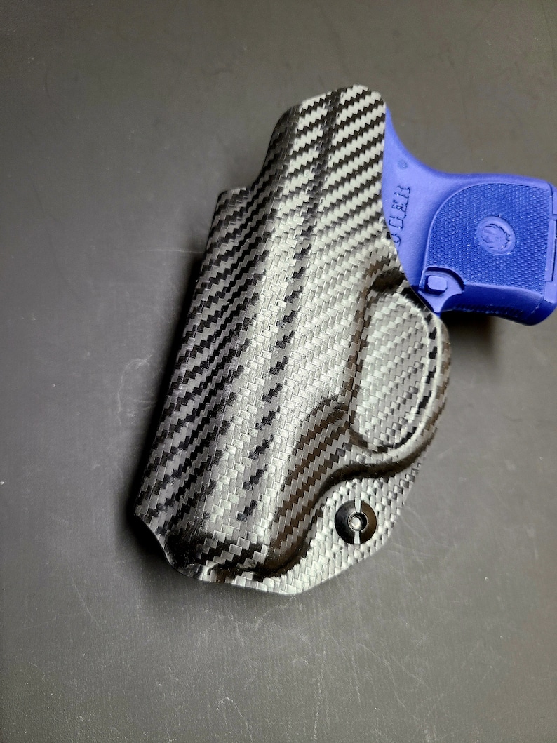 May include: A black carbon fiber holster for a small handgun. The holster has a single screw on the bottom and the text "CLEAR" is visible on the side.