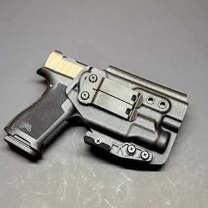 May include: A black polymer gun holster with a black metal clip and a black handgun inside. The holster has a textured surface and a small American flag logo on the side.