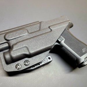 May include: A black polymer holster with a textured surface and a black handgun inside. The holster has a belt clip and three screws on the bottom.